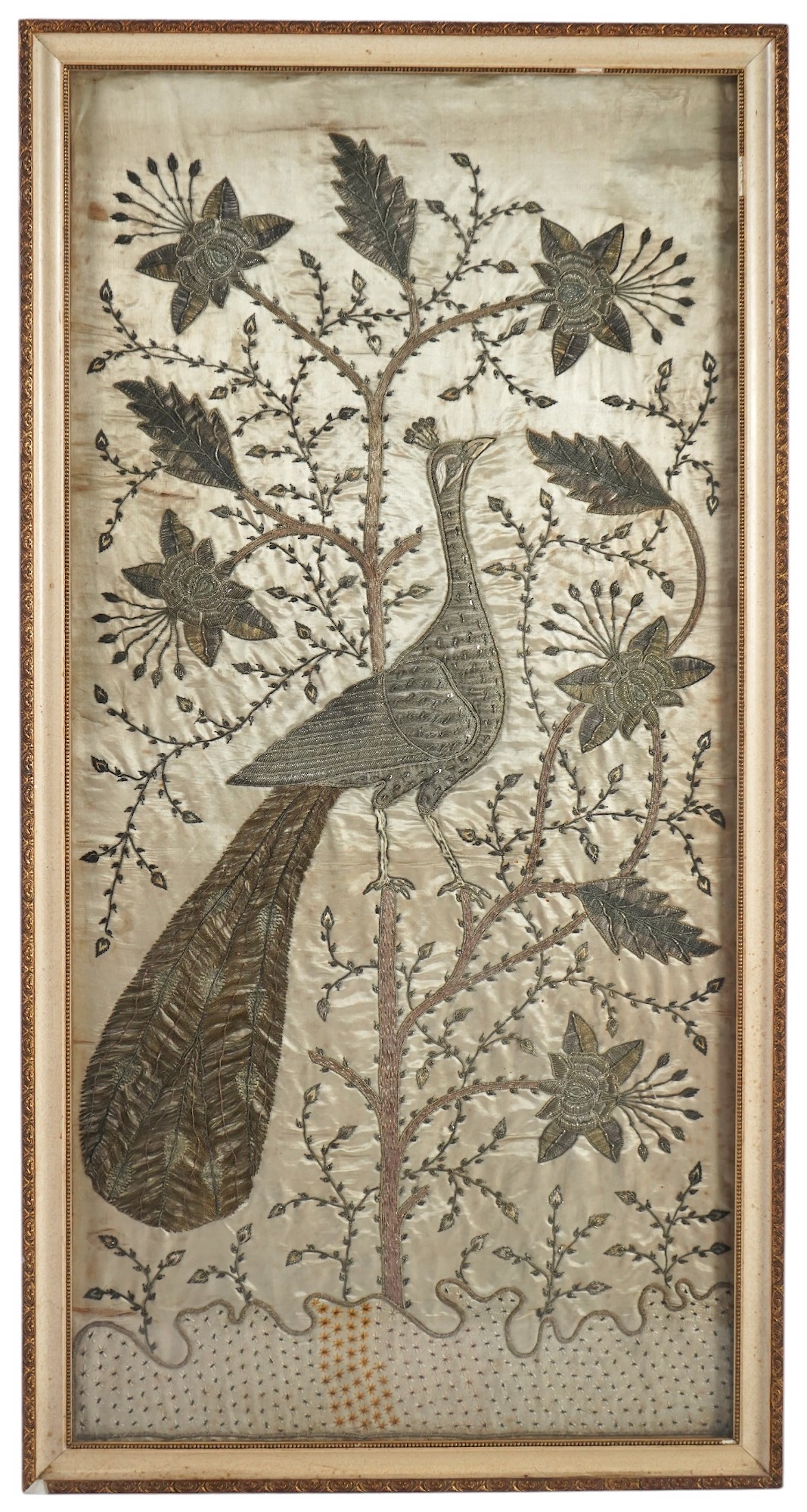 An Indian late 19th century silver metallic thread embroidery of a peacock in a flowering tree, embroidered on silk, parts of the embroidery worked in relief like stump work, 107cm x 52cm, framed. Condition - silk backin